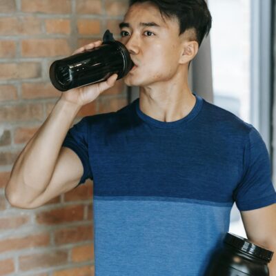 Fit young man drinking water after workout indoors holding protein bottle.