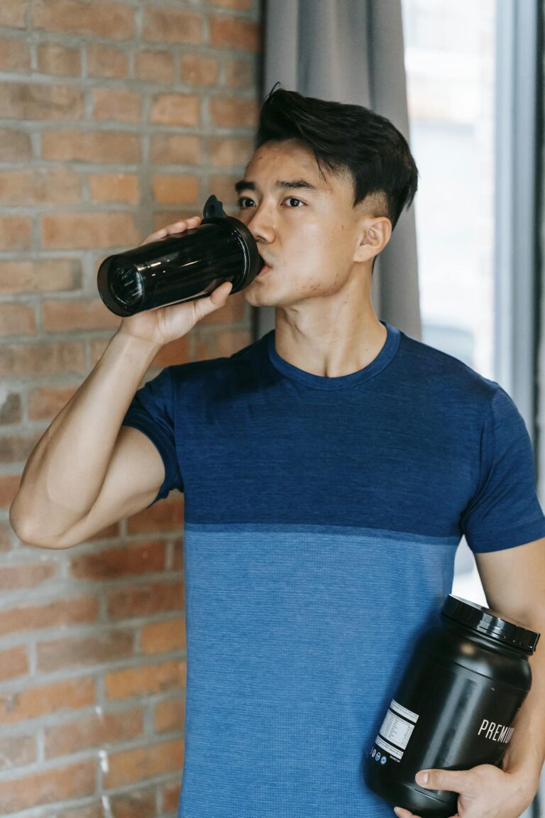 Fit young man drinking water after workout indoors holding protein bottle.