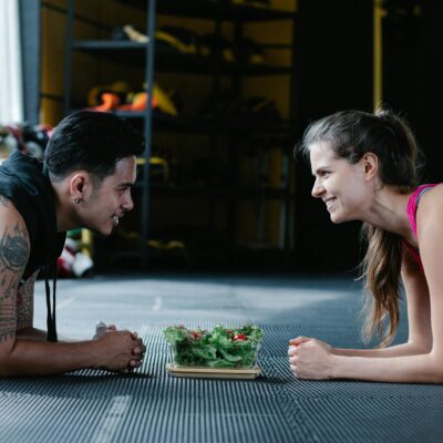 Two people planking face-to-face in a gym with a salad between them, smiling and engaging in a fitness activity.