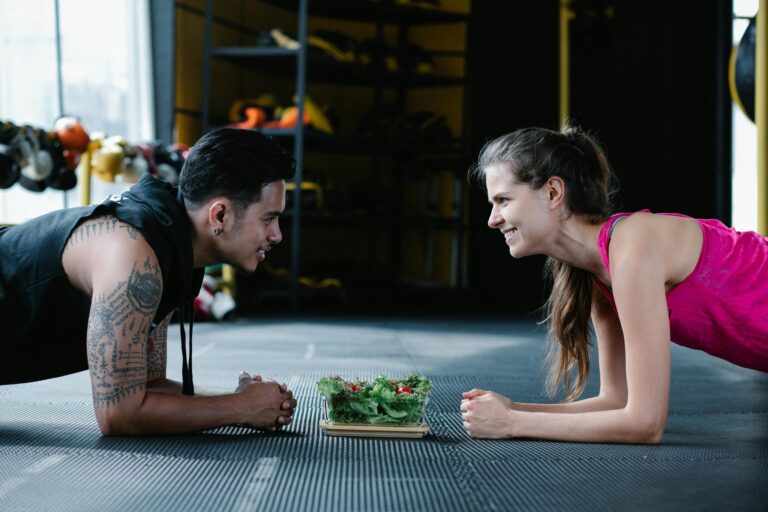 Two people planking face-to-face in a gym with a salad between them, smiling and engaging in a fitness activity.